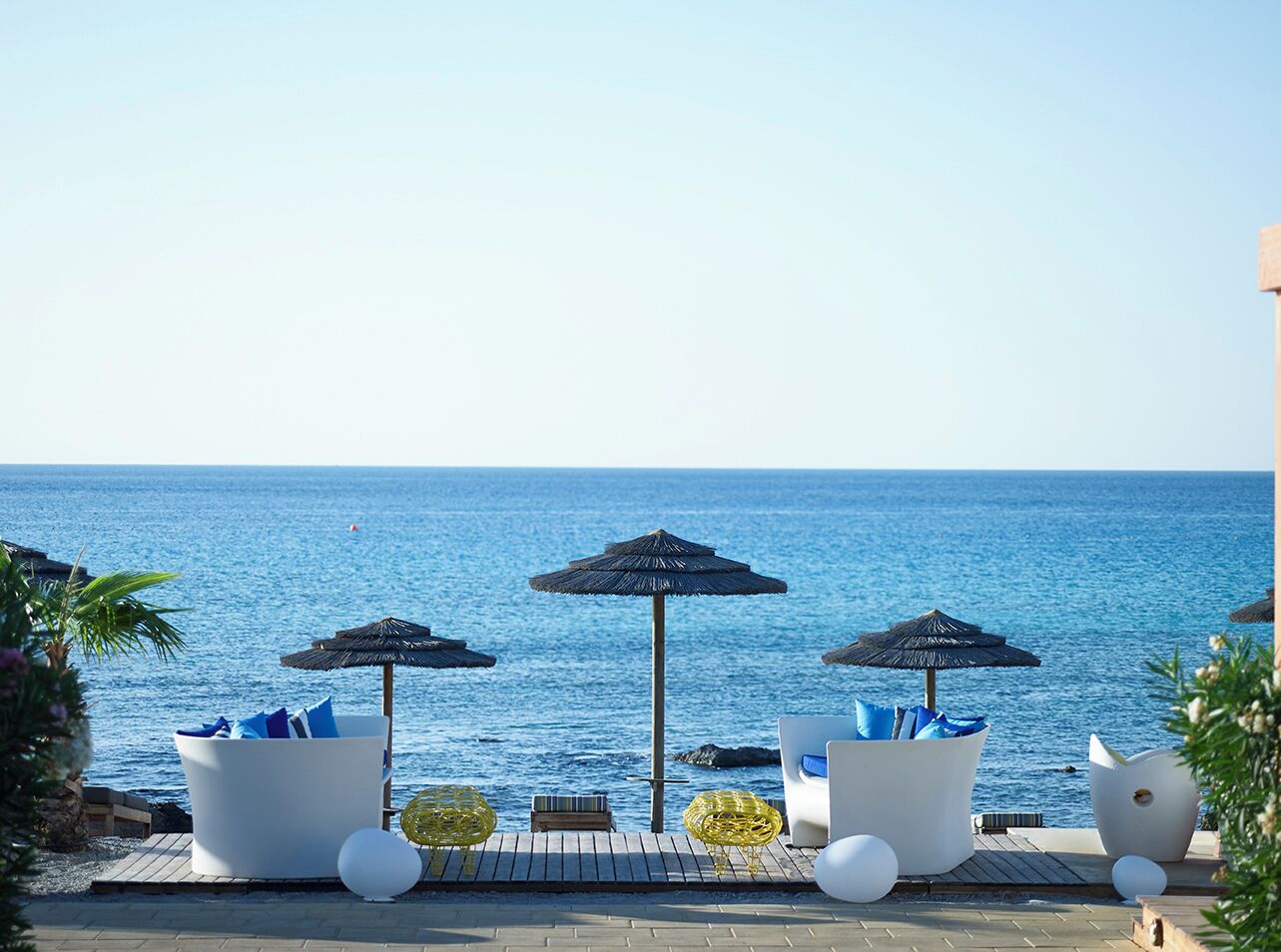 A seaside lounge area with three white lounge chairs, each with a blue cushion, and a thatched umbrella.