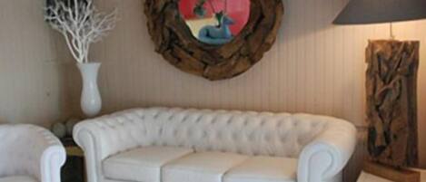 A white upholstered sofa in a room with a decorative wooden frame on the wall, a white vase with branches, and a lamp on a side table.
