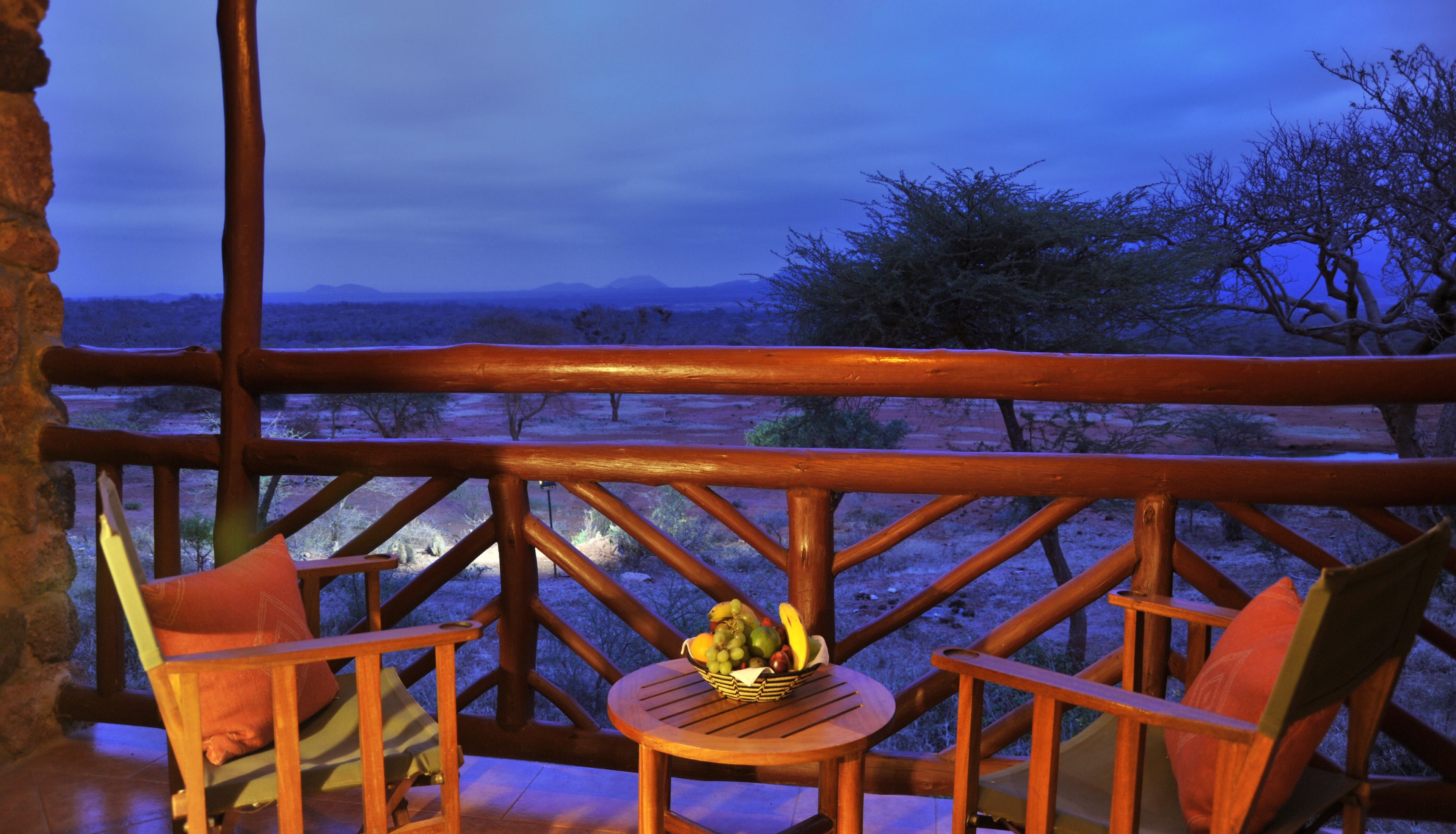 Una terraza de madera con dos sillas y una mesa, con vista a un paisaje desértico de vegetación escasa y montañas lejanas.