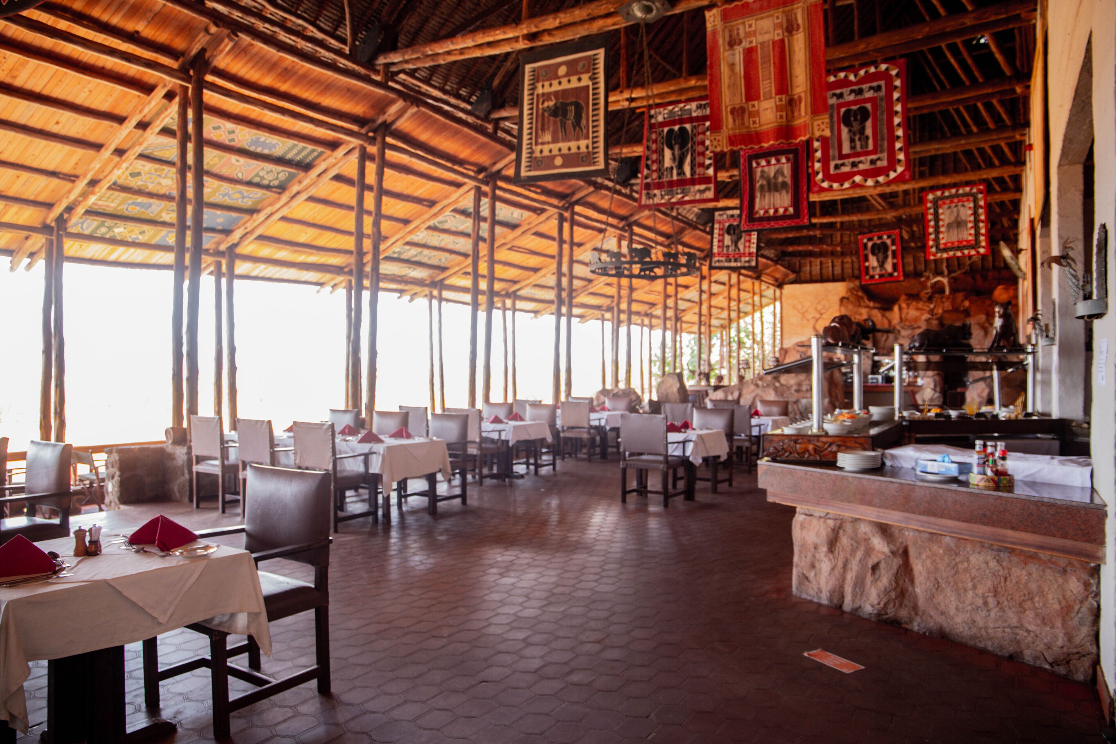 Un restaurante con techos altos, azulejos decorativos en el techo y una zona de bufé con mostrador de piedra. Las mesas están dispuestas con manteles blancos y servilletas rojas.