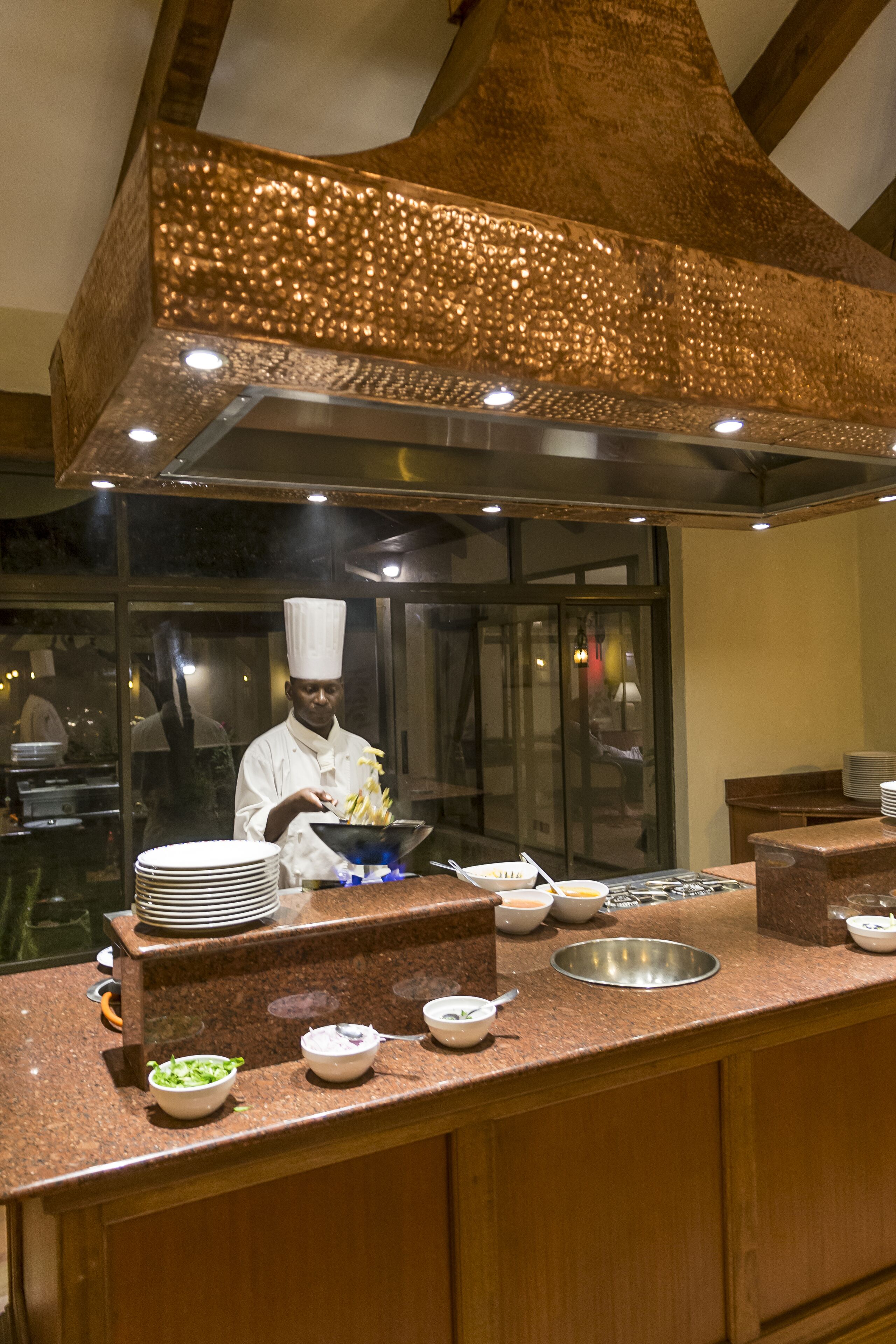 A chef preparing food in a well-lit kitchen with a large copper hood and stainless steel appliances.