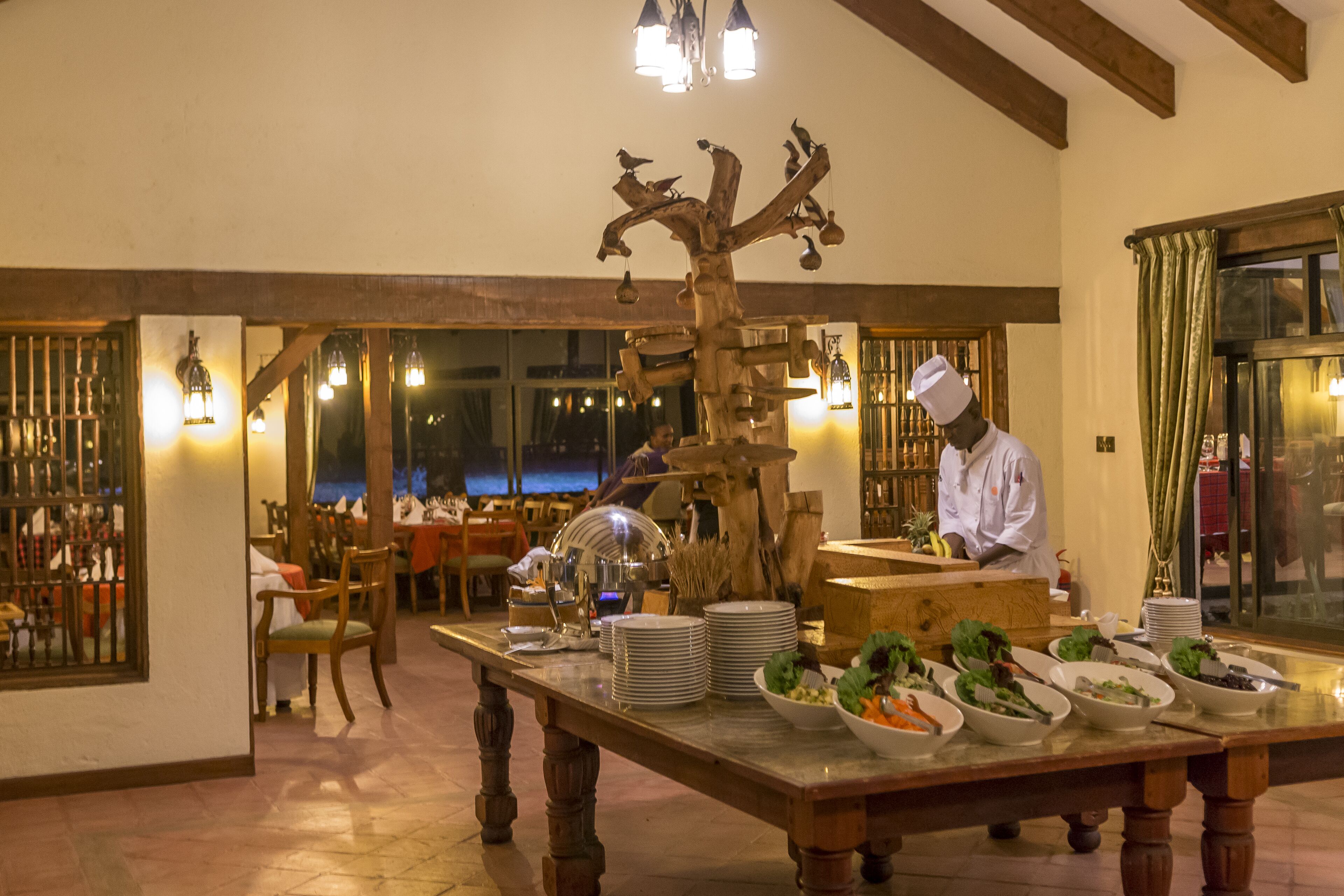 A restaurant with a long wooden table set for a meal, a chef preparing food, and a decorative tree sculpture.