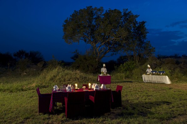 Un espace de restauration en plein air avec des tables et des chaises, un chef qui prépare les plats et un grand arbre en arrière-plan.