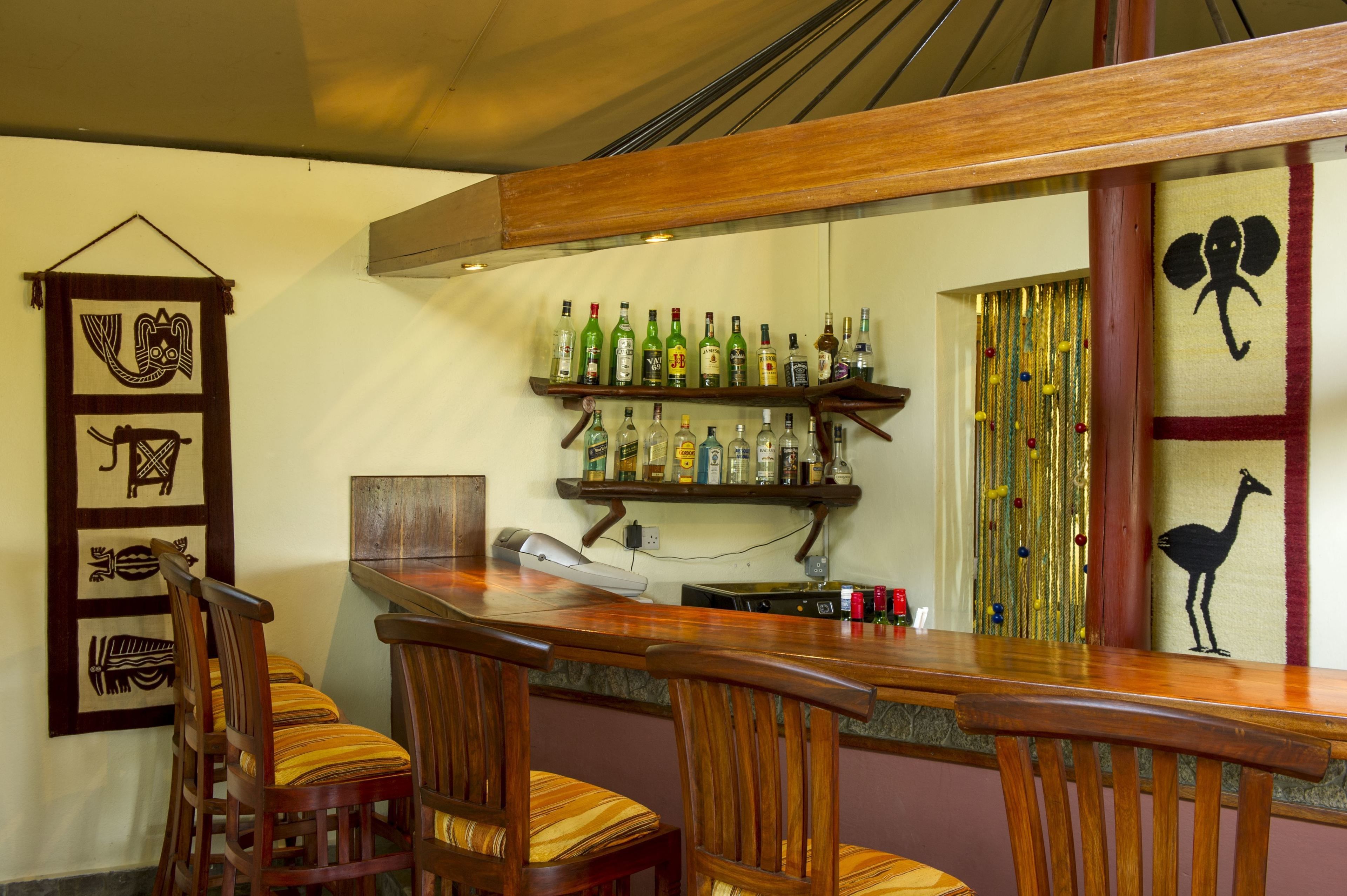 A bar area with wooden chairs and a long counter. There are shelves with bottles and a decorative wall hanging.