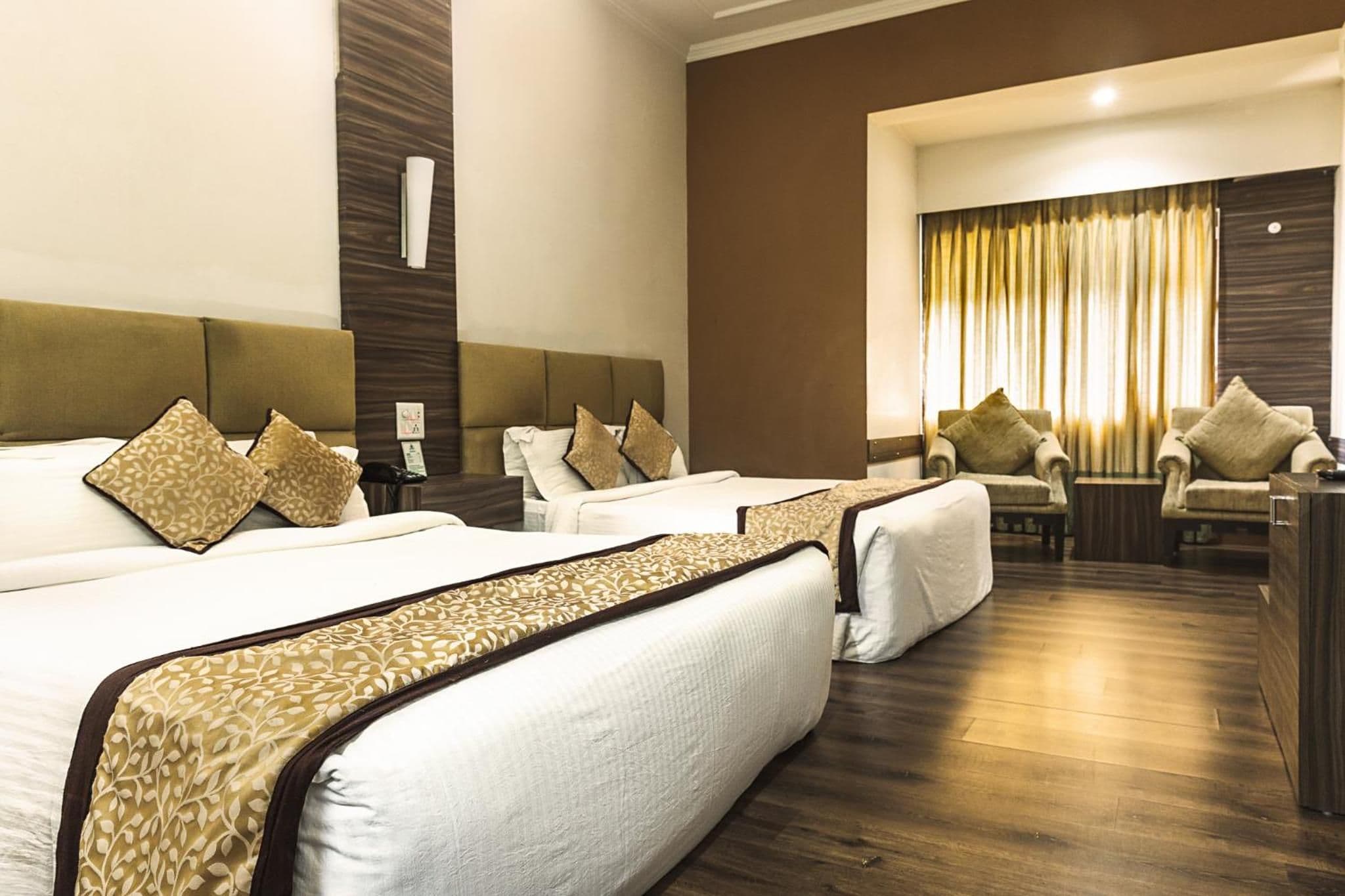 A hotel room with three beds, a seating area with armchairs, and a large window with curtains.