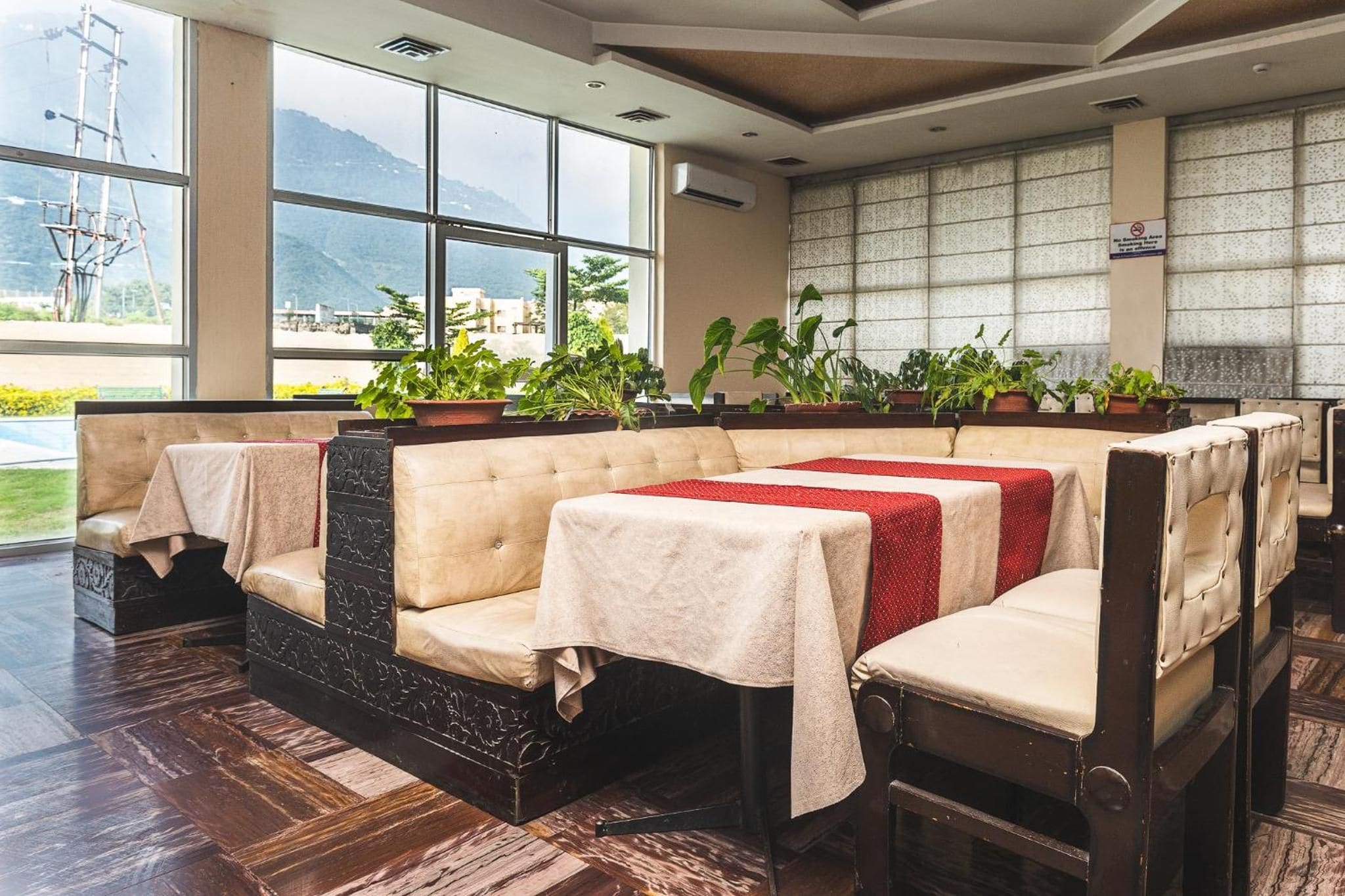 A restaurant with cushioned seating, tables with tablecloths, potted plants, and a view of mountains through large windows.