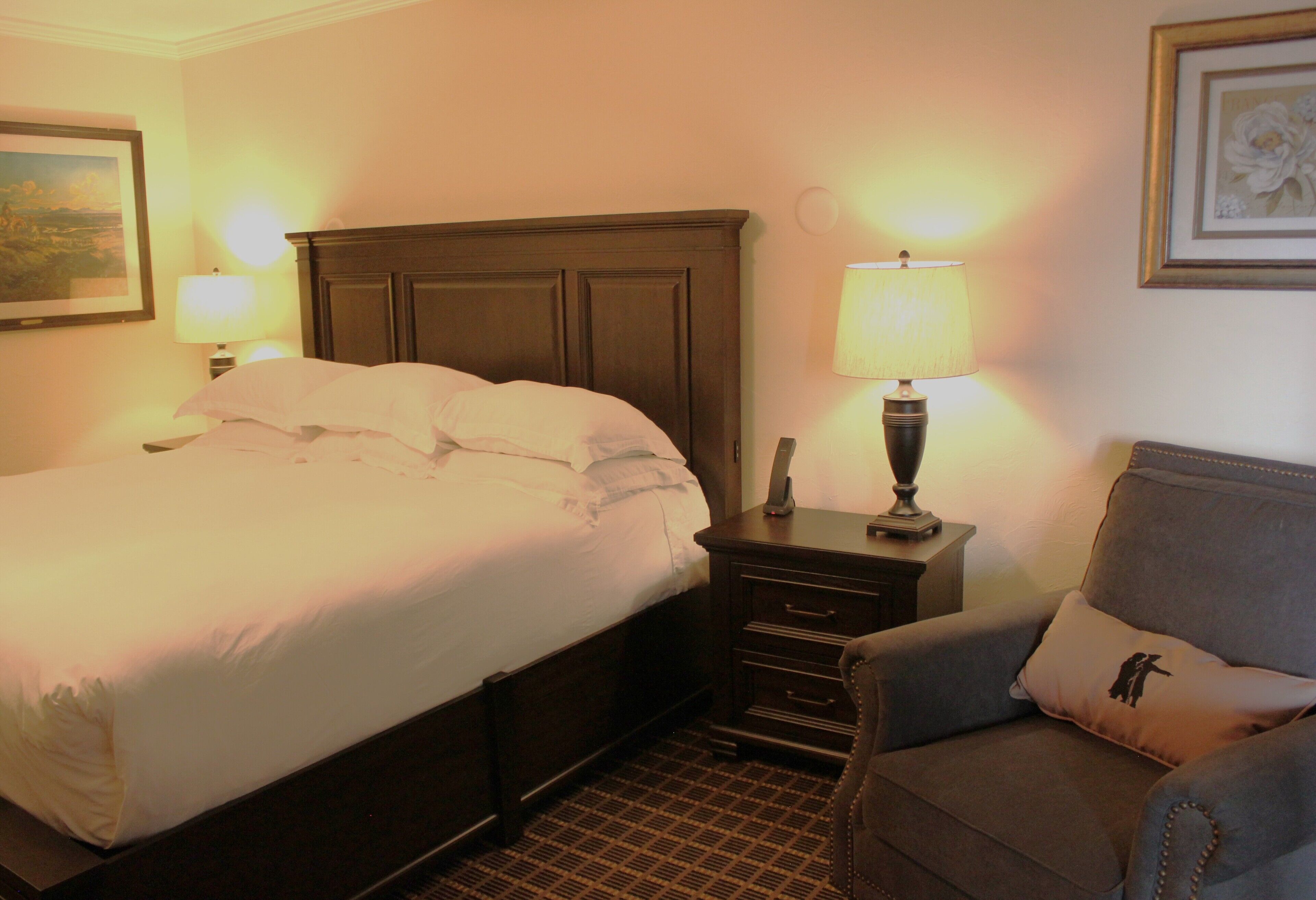 A hotel room with a large bed, a nightstand with a lamp, a chair, and a framed picture on the wall.
