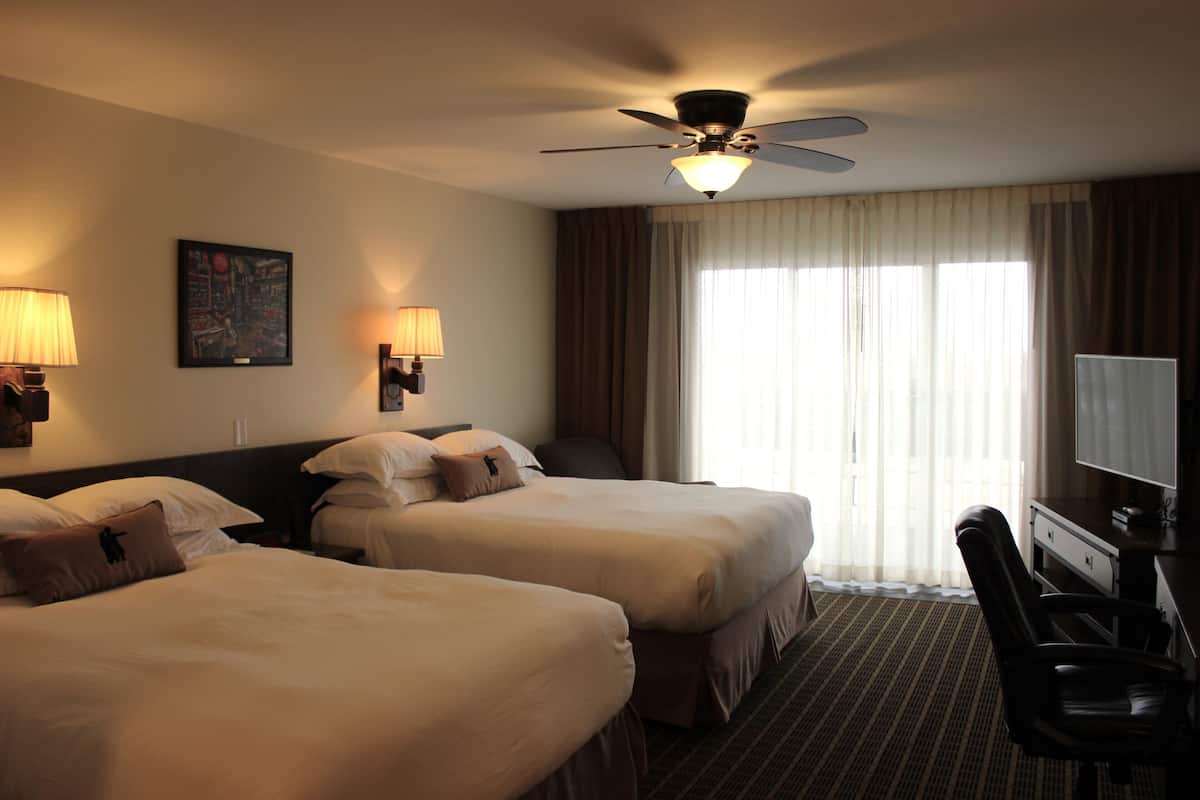 A hotel room with two beds, a desk, a chair, a ceiling fan, and a large window with curtains.