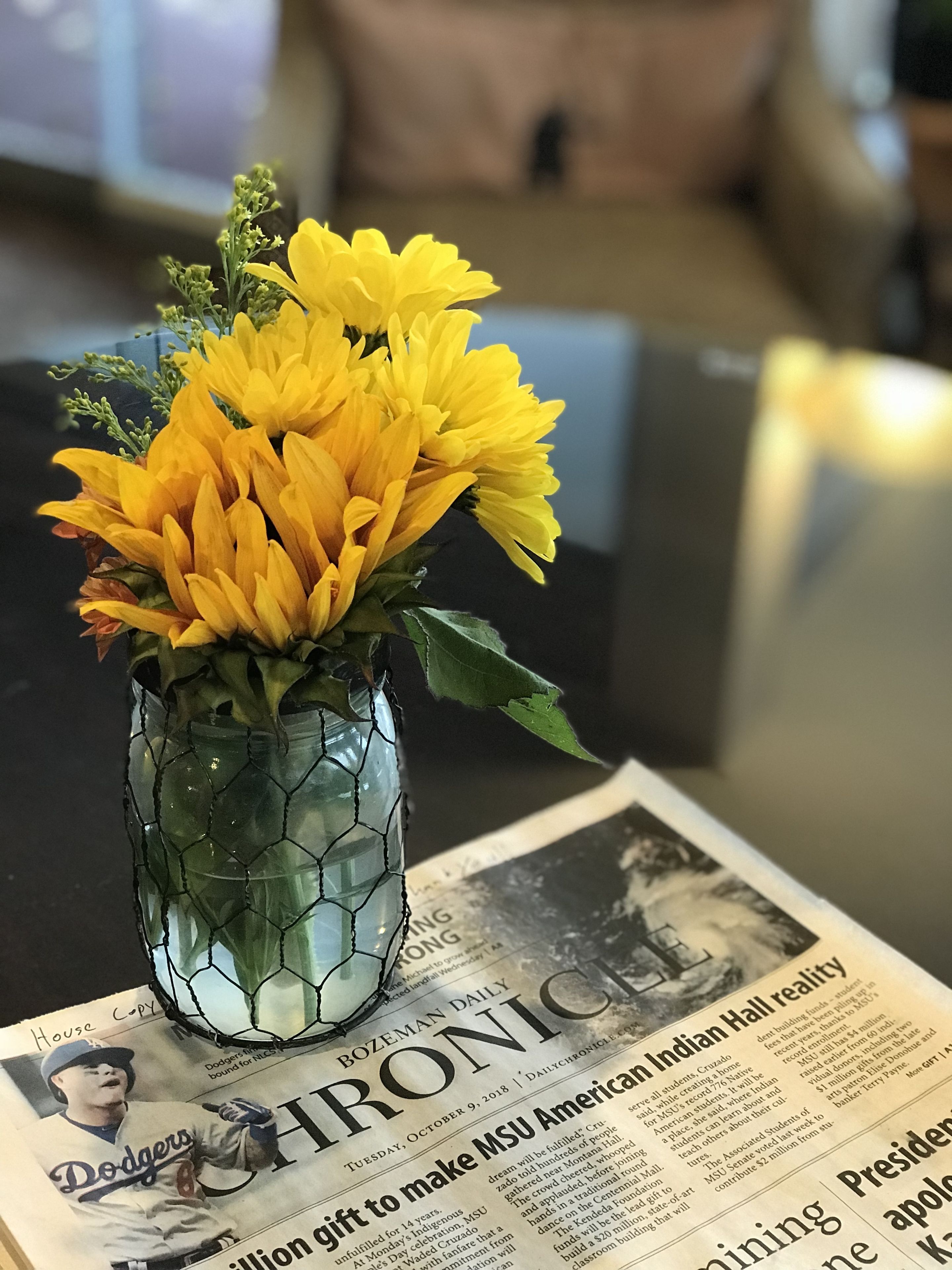 A vase with yellow flowers on a table, placed on a newspaper with a sports section.