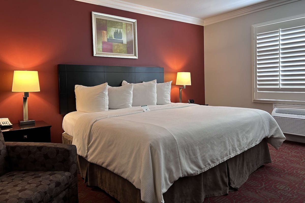 A hotel room with a large bed, two bedside lamps, a painting on the wall, and a window with blinds.