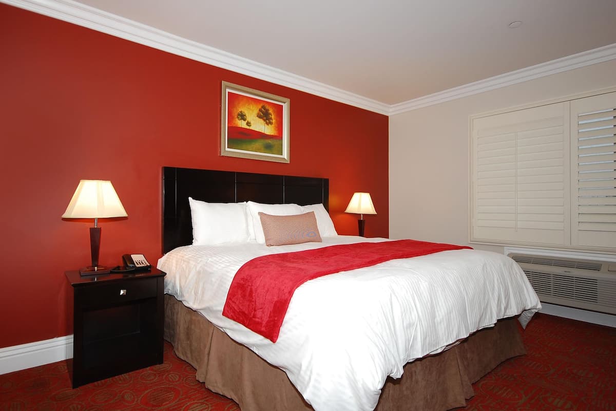 A hotel room with a large bed, two bedside lamps, a painting on the wall, a telephone, and a window with blinds.