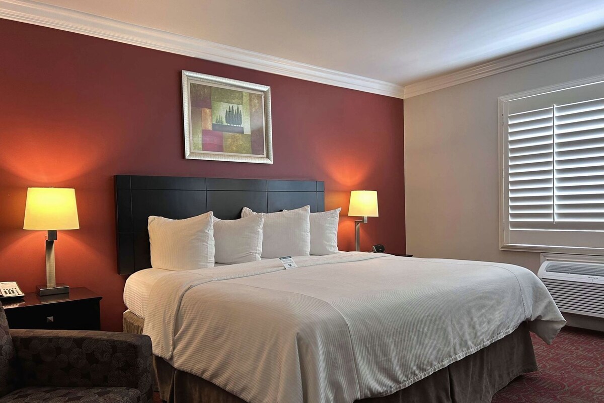 A hotel room with a large bed, two bedside lamps, a chair, a painting on the wall, and a window with blinds.
