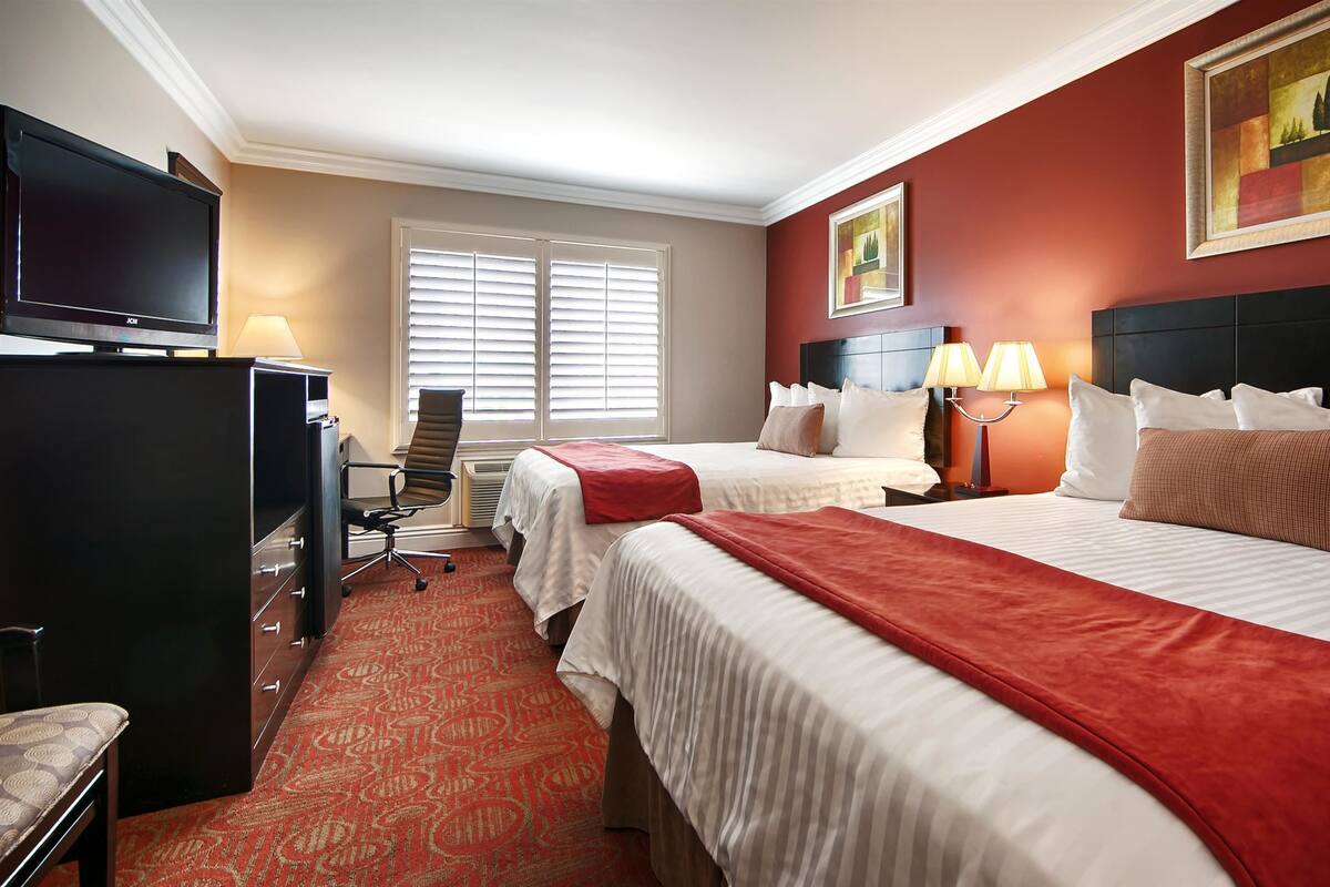 A hotel room with two beds, a desk, a TV, and a window with blinds.