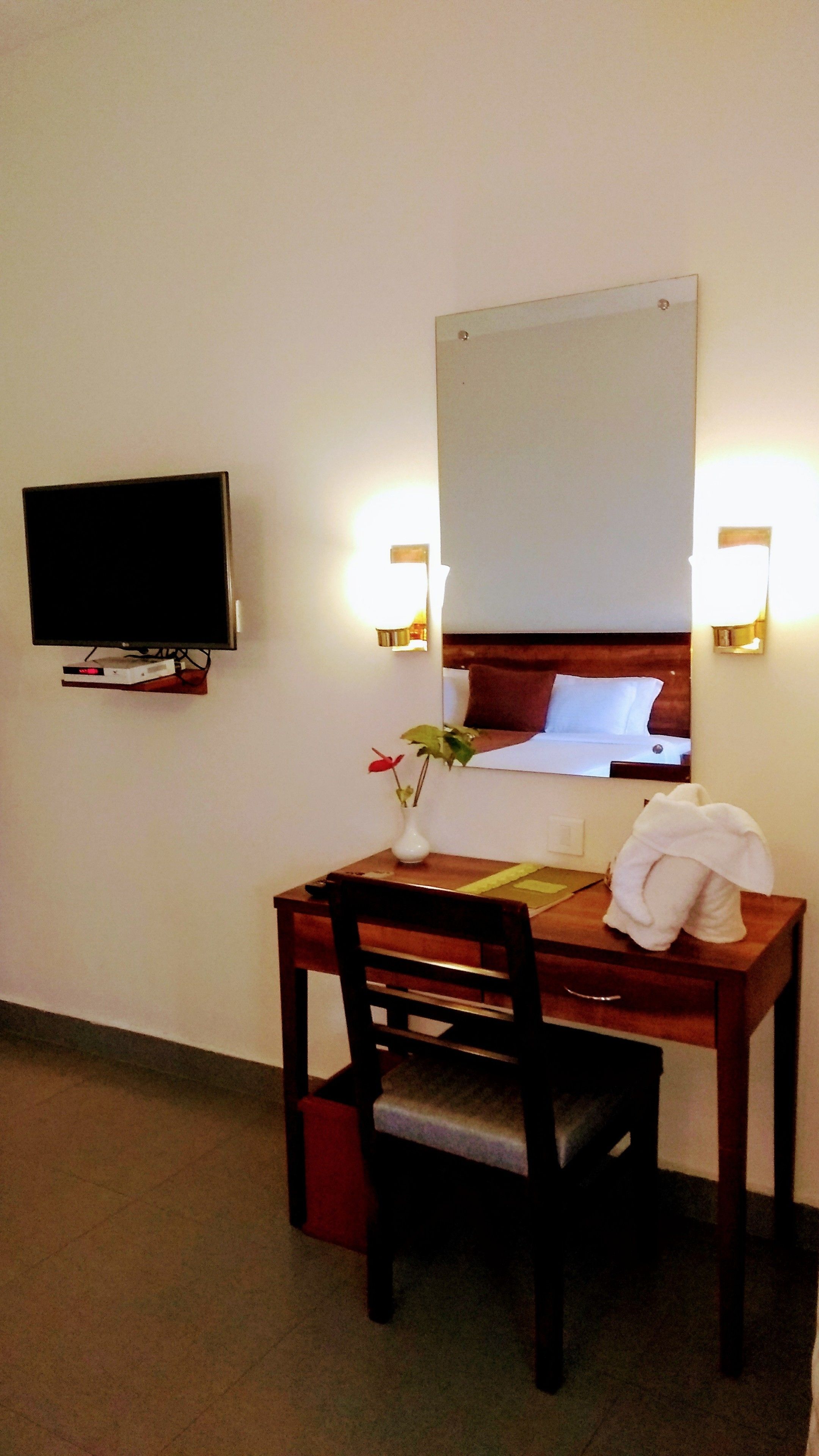 Habitación de hotel con un escritorio de madera, una silla, un televisor montado en la pared, un espejo y una toalla blanca sobre la silla.