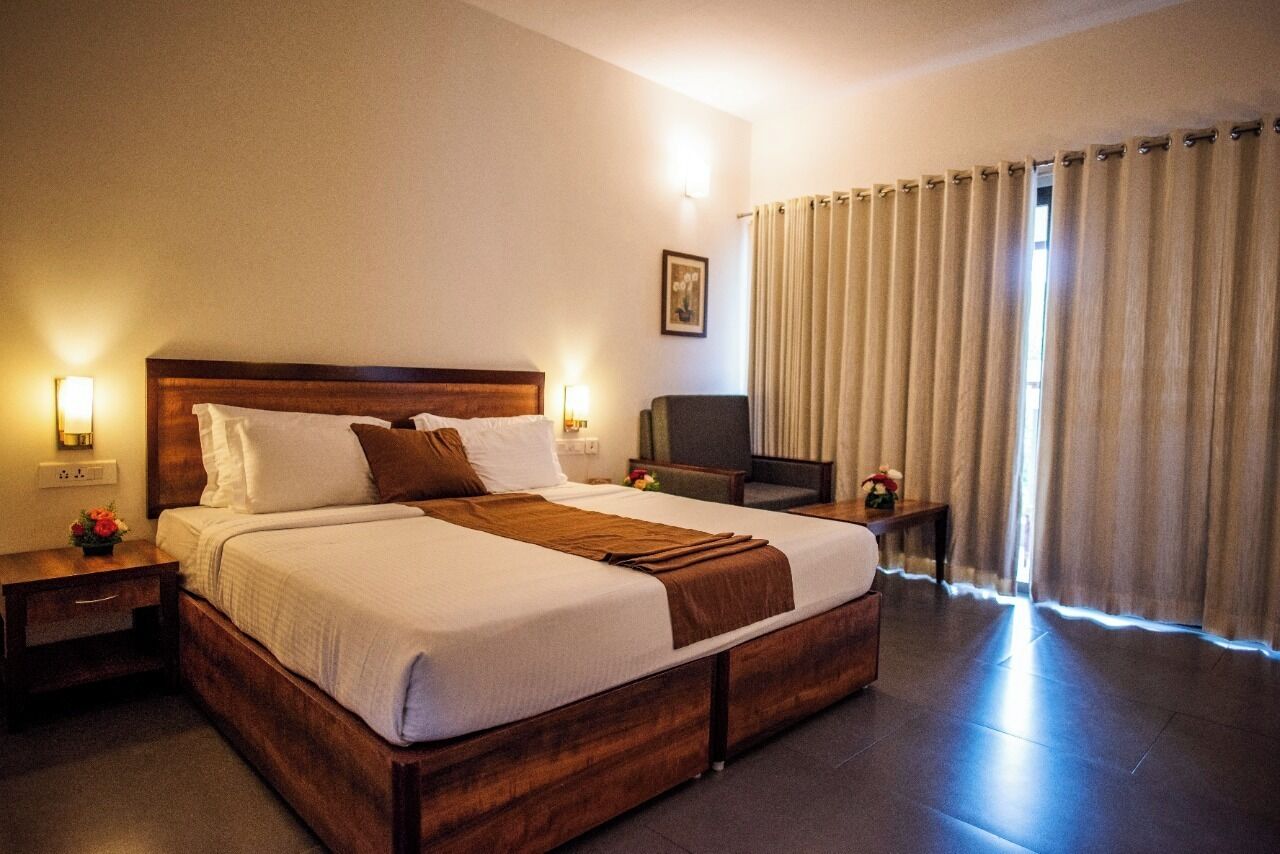 Habitación de hotel con una cama grande, cabecera de madera, dos mesitas de noche con lámparas, un televisor y una ventana con cortinas.