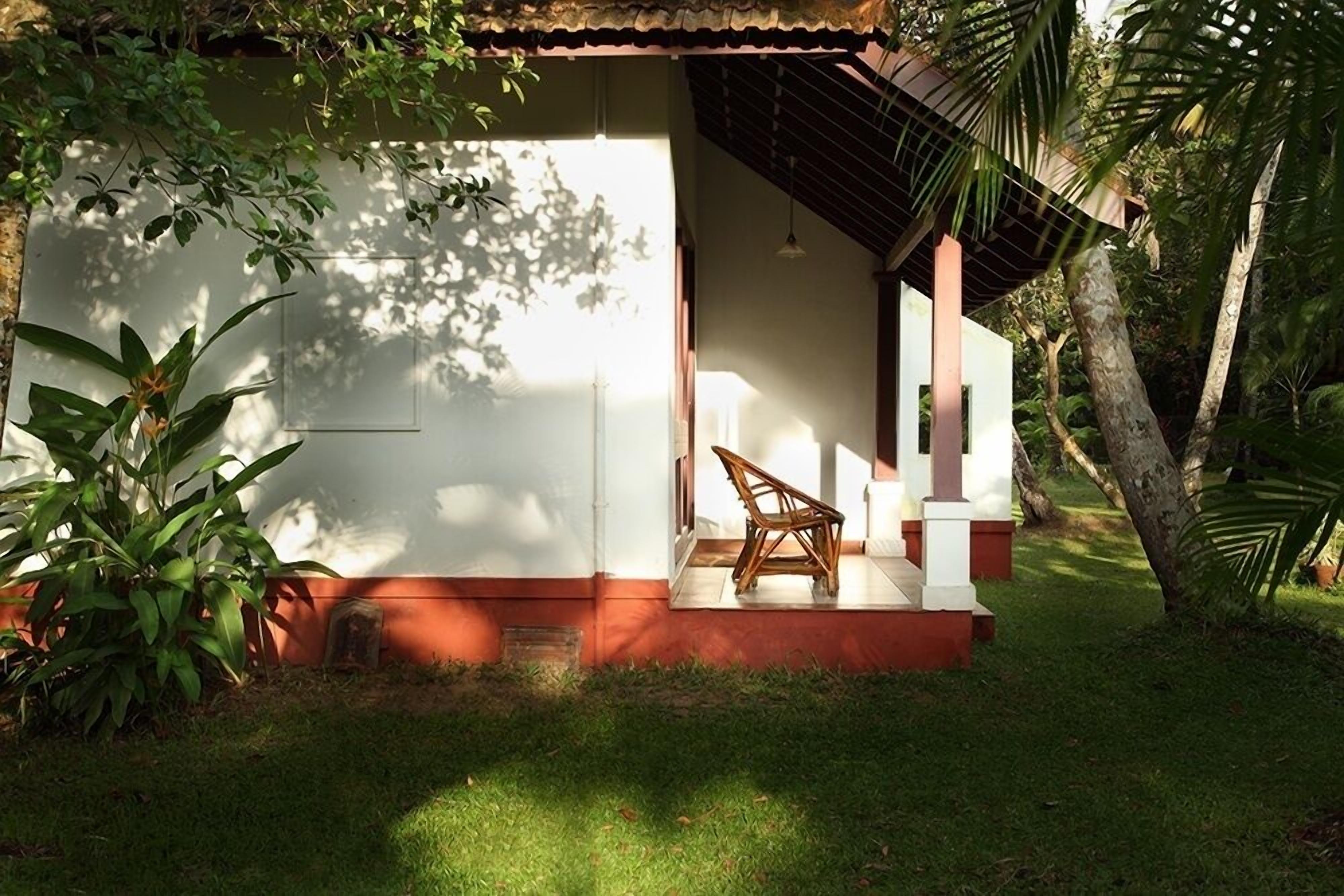 A small, single-story building with a red and white exterior, a covered porch with a chair, and lush greenery surrounding it.
