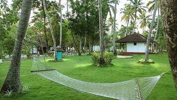 A hammock in a lush, green garden with palm trees and a small white building.
