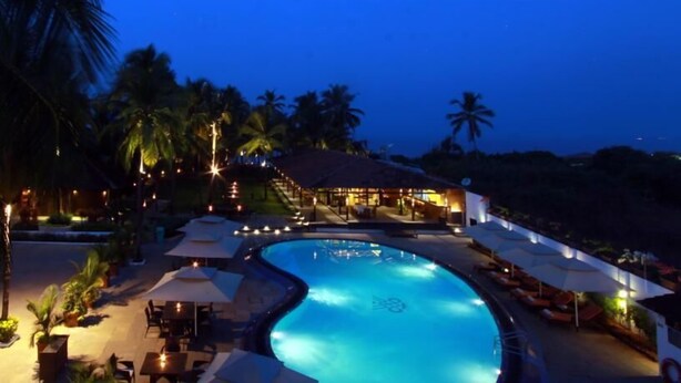 Ein Poolbereich eines Resorts mit Liegestühlen und Sonnenschirmen, umgeben von Palmen und einem Gebäude mit einem Reetdach.