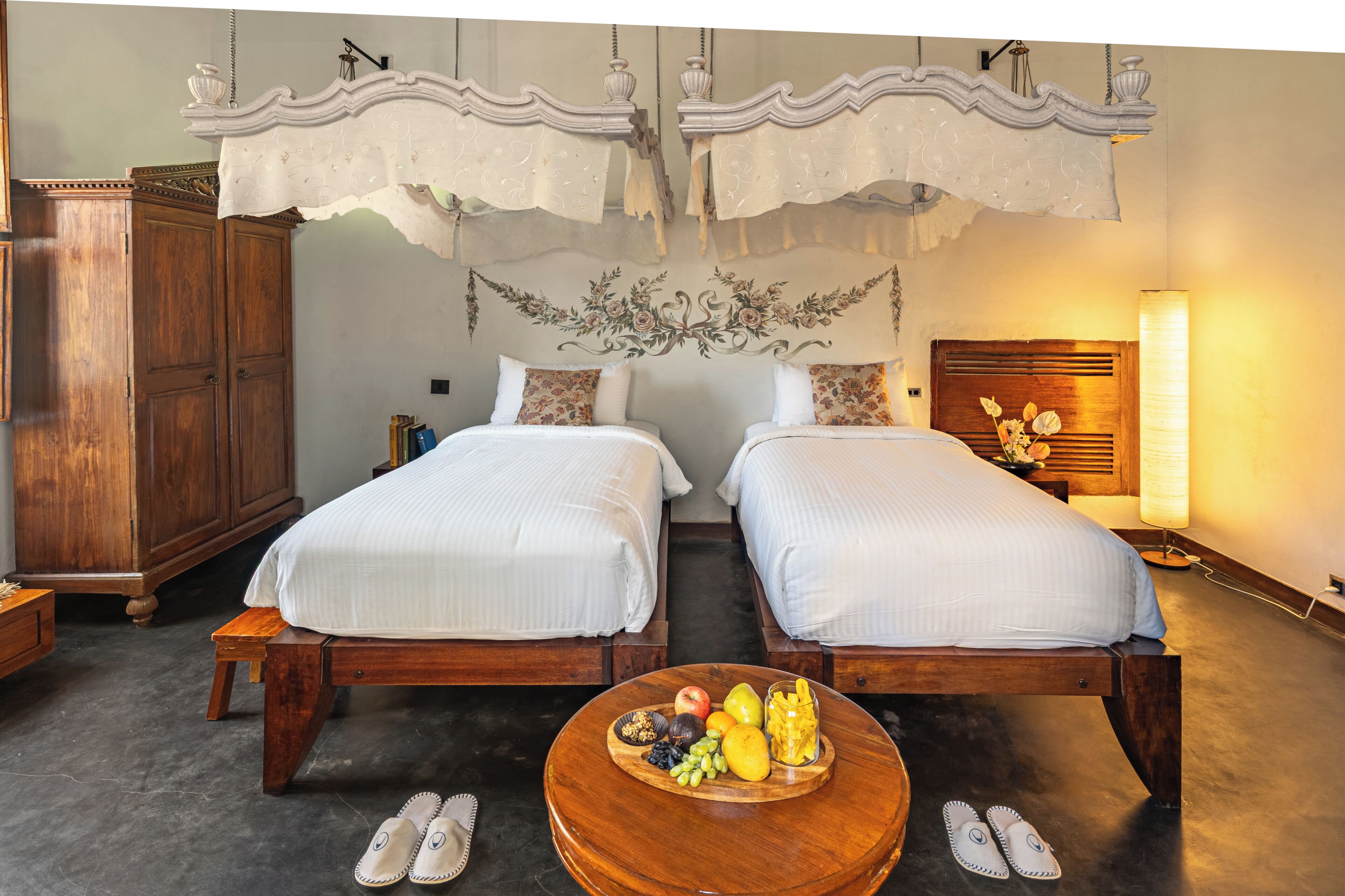 Kahden hengen huone, jossa on kaksi sänkyä, puinen vaatekaappi, pyöreä puinen pöytä, jonka päällä on hedelmiä ja tossut, sekä katosta roikkuva verho.