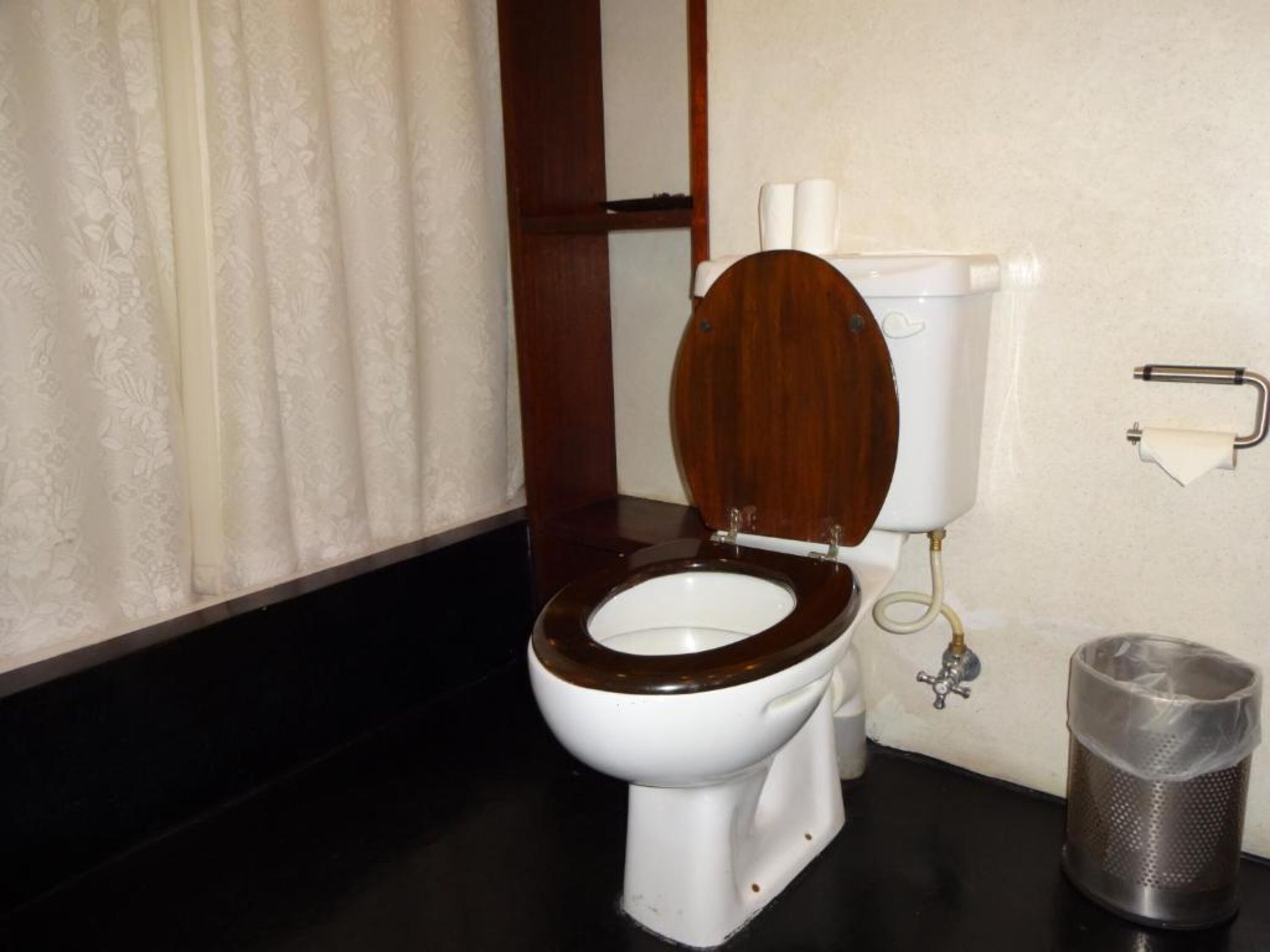 A standard toilet with a wooden seat and lid, a white tank, and a metal trash can.