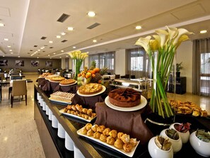 A buffet setup with various desserts and pastries, a buffet line with white plates, and a decorative arrangement with flowers and fruits. - Eurostars Roma Aeterna (Rome)