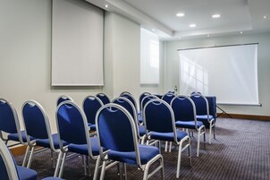 Konferenssihuone, jossa on riveissä sinisiä tuoleja, valkoinen projektorikangas ja matolla peitetty lattia.