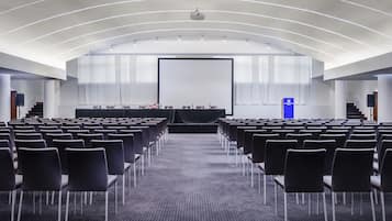 Un salón de conferencias con filas de sillas, un escenario con proyector y un atril.