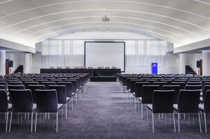 Una sala conferenze con file di sedie, un palco con un proiettore e un podio.