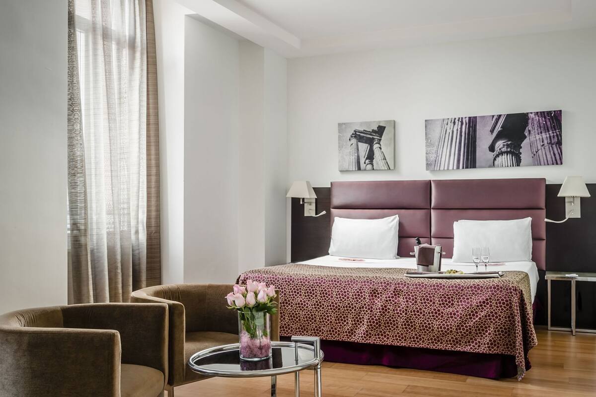 A hotel room with a bed, two chairs, a small table with a vase of flowers, and artwork on the wall.