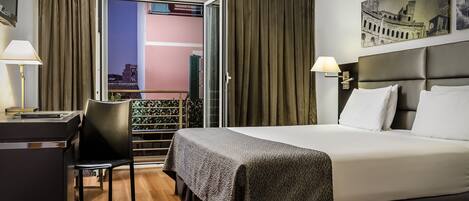 A hotel room with a bed, desk, chair, and a balcony with a view of a building.