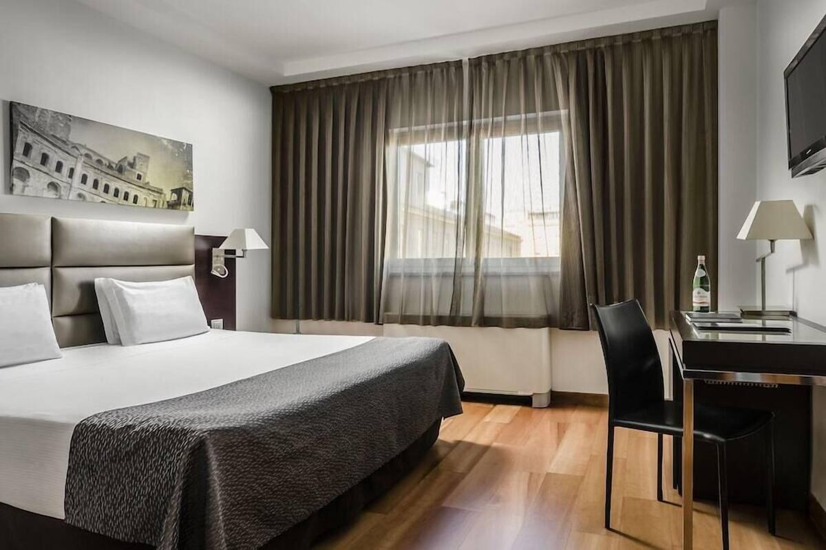A hotel room with a large bed, a desk with a chair, a television, and a window with curtains.