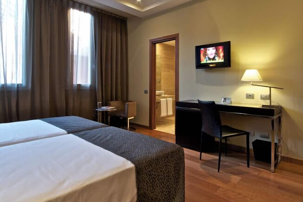 A hotel room with two beds, a desk, a chair, a TV, and a lamp.