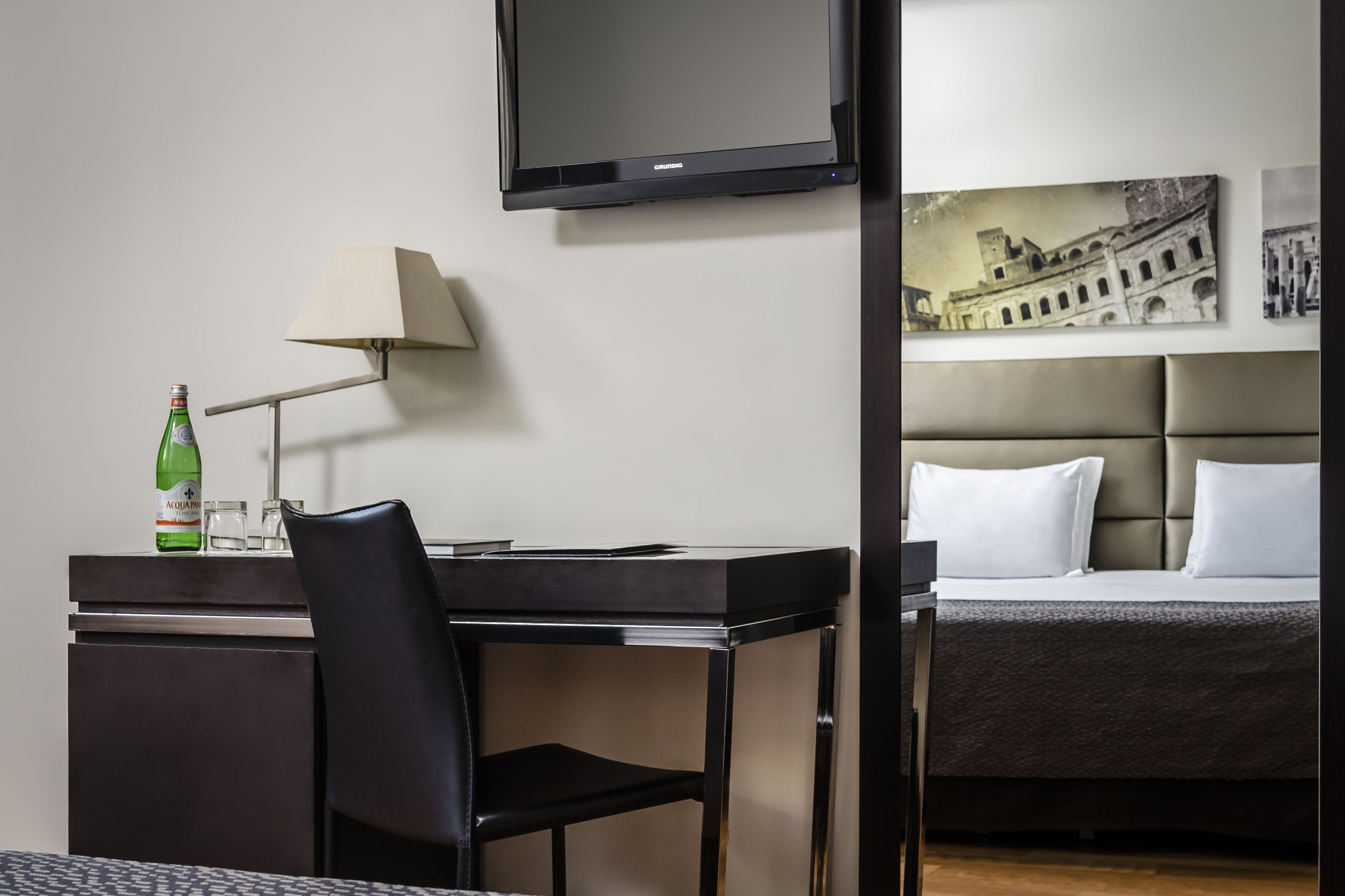 Quarto de hotel com uma cama, uma escrivaninha com cadeira, uma luminária e uma televisão na parede.