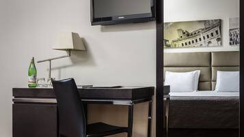 Quarto de hotel com uma cama, uma escrivaninha com cadeira, uma luminária e uma televisão na parede.