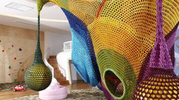 A colorful knitted canopy hanging in a room with a wooden floor, a white chair, and a climbing wall in the background.