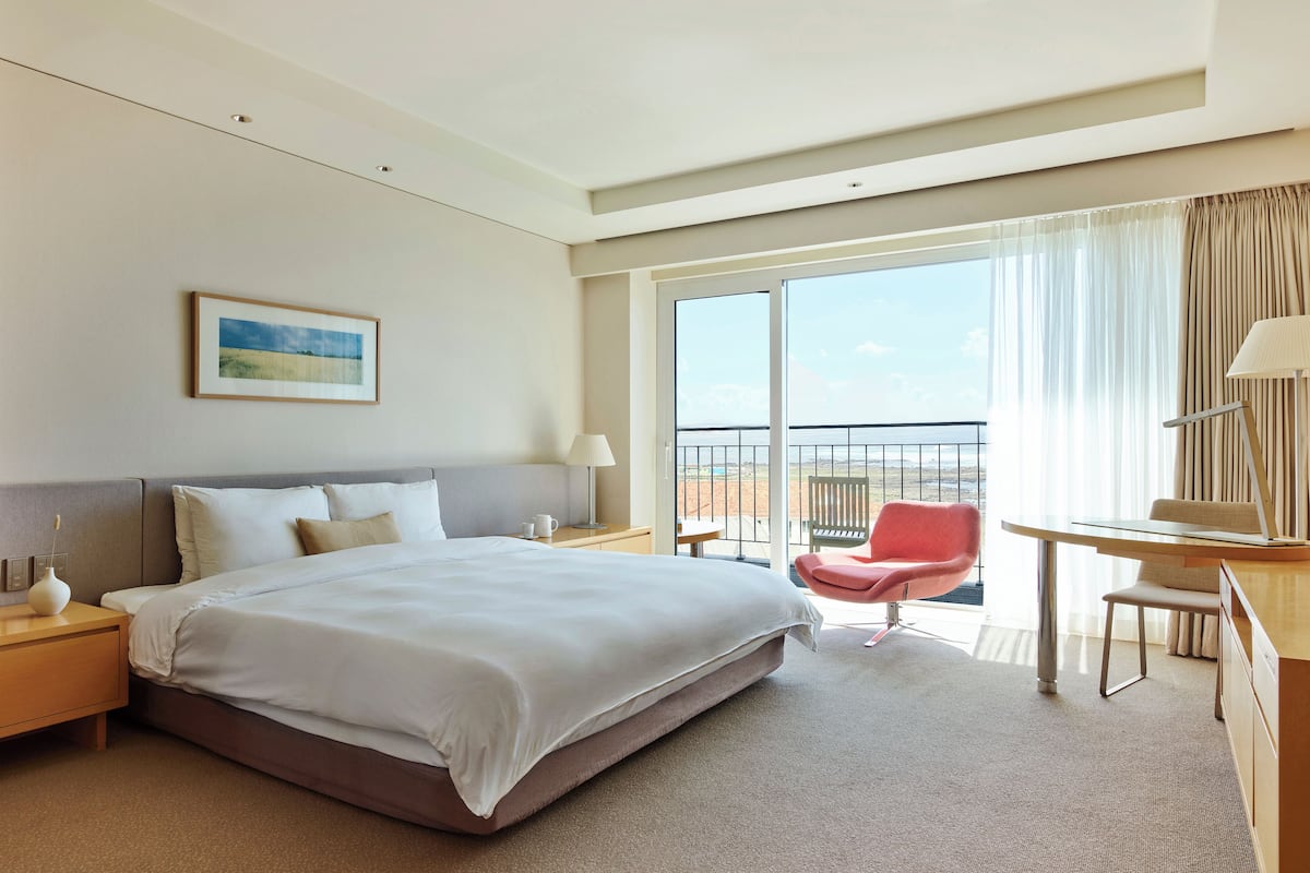 Une chambre d’hôtel avec un grand lit, un bureau, une chaise, une table de chevet, une lampe, un tableau au mur et un balcon avec vue.