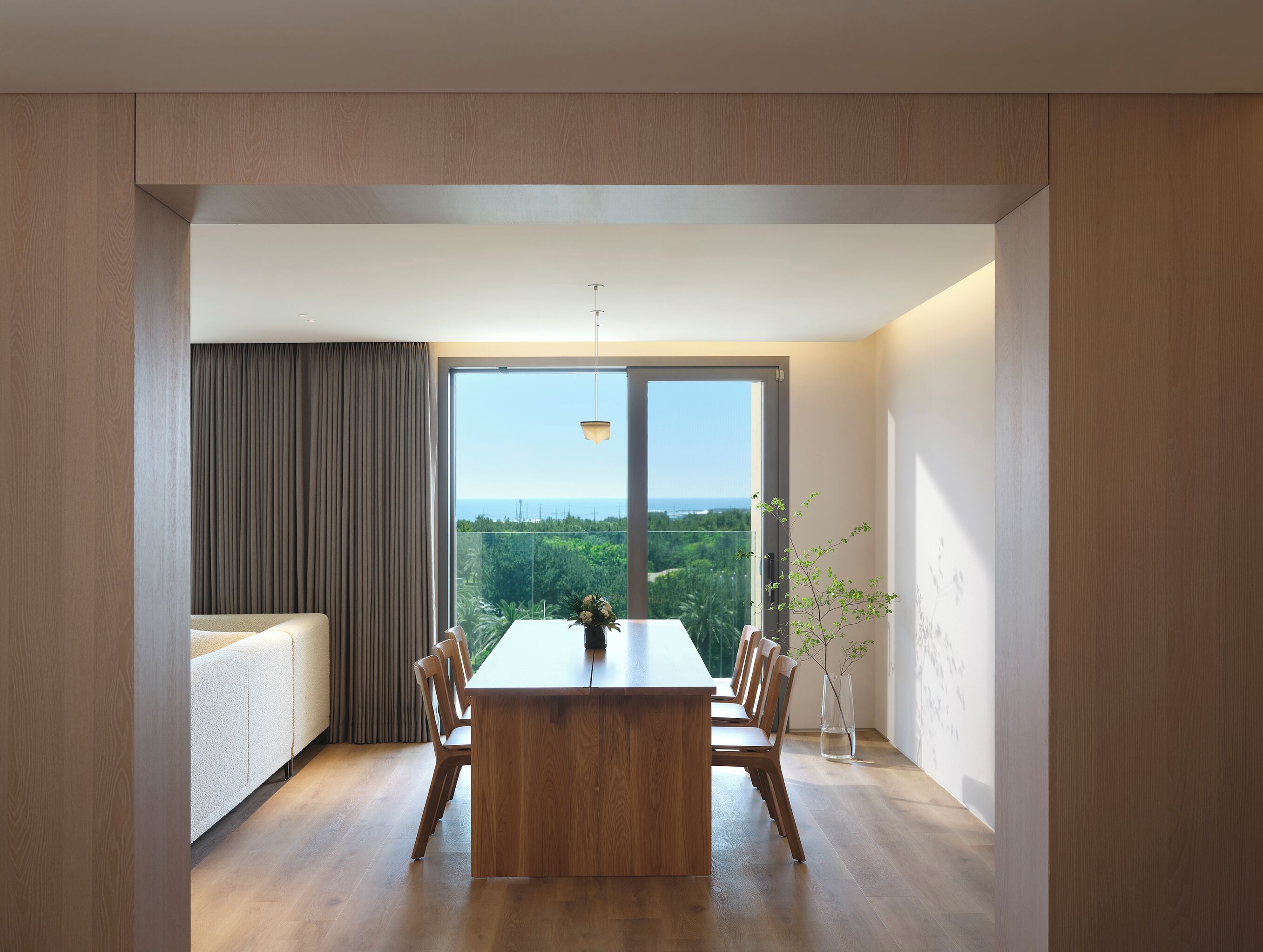 A modern dining area with a wooden table and chairs, a large window with a view of greenery and a distant landscape, and a cozy seating area with a sofa and curtains.