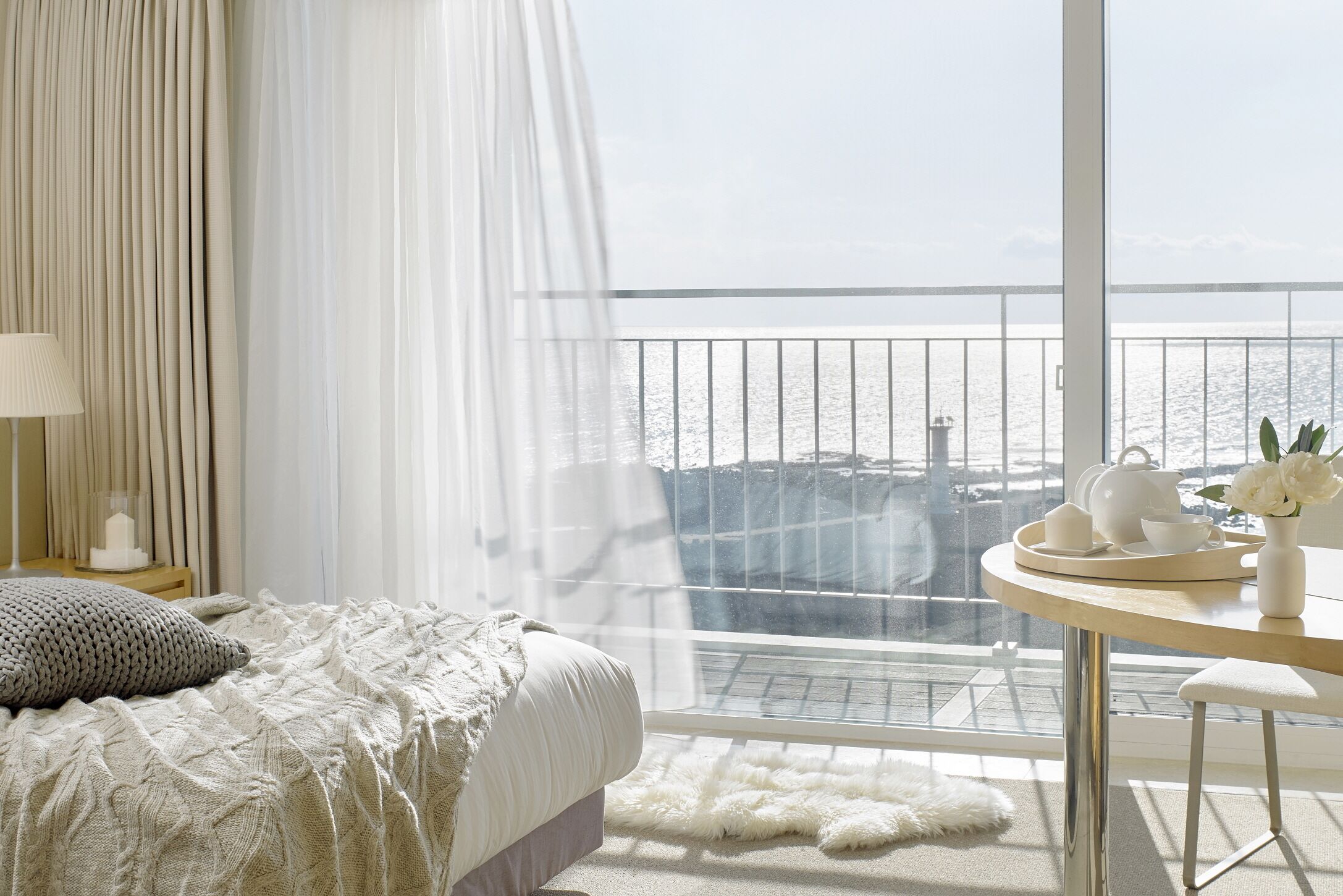 A bedroom with a large window overlooking a balcony with a view of the sea.