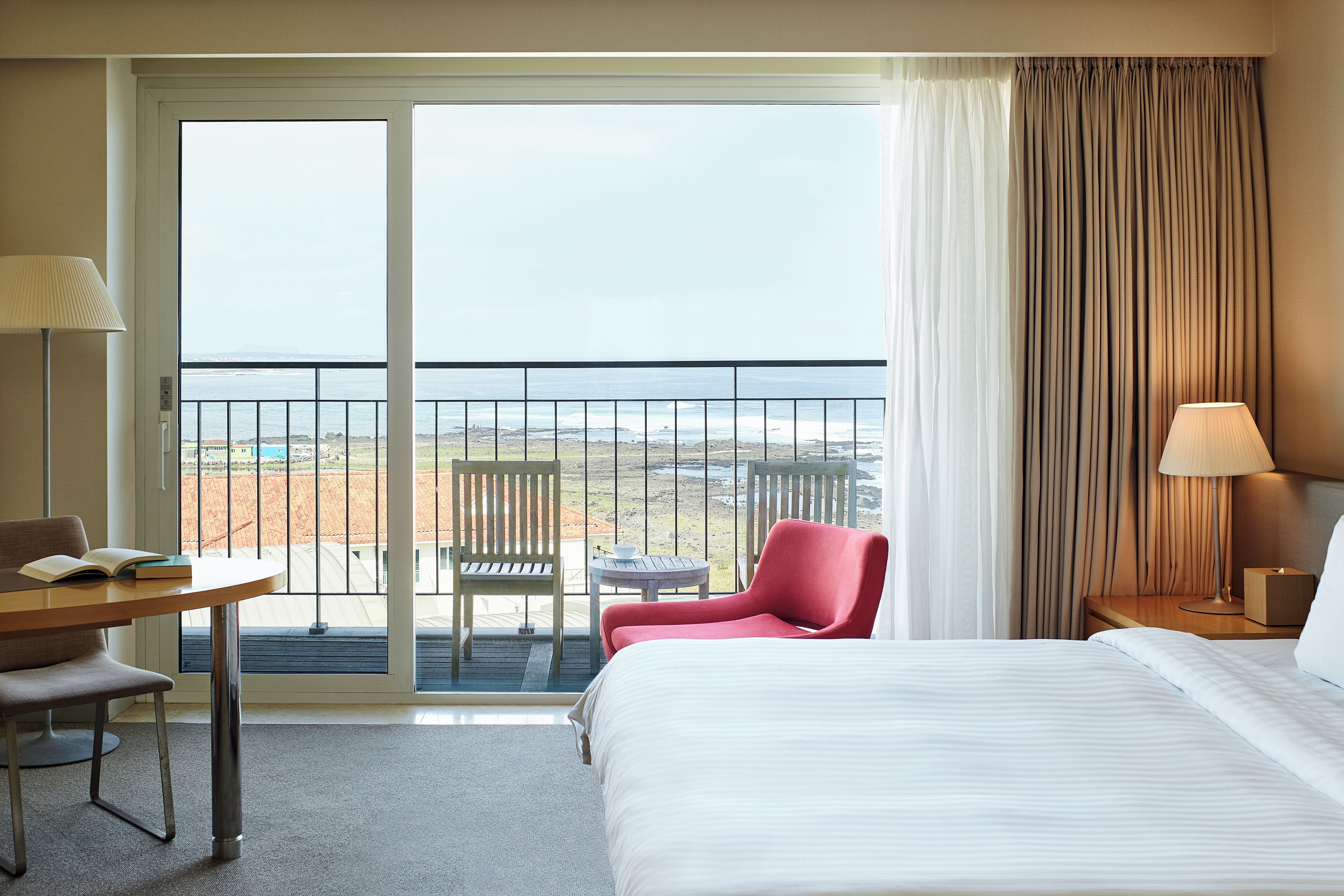 Habitación de hotel con cama, escritorio, silla, mesa y vista al mar desde un balcón.