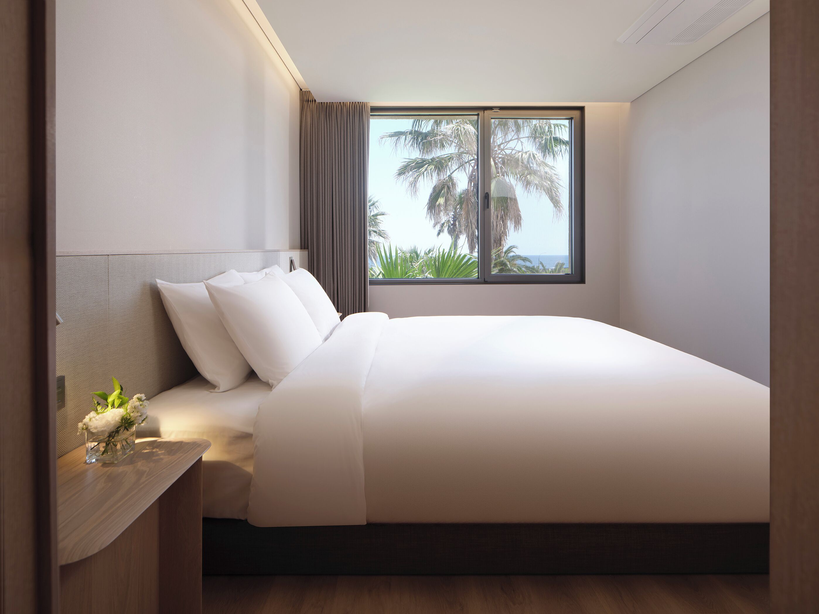 A modern hotel room with a large bed, a nightstand, a window with a view of palm trees, and a headboard.