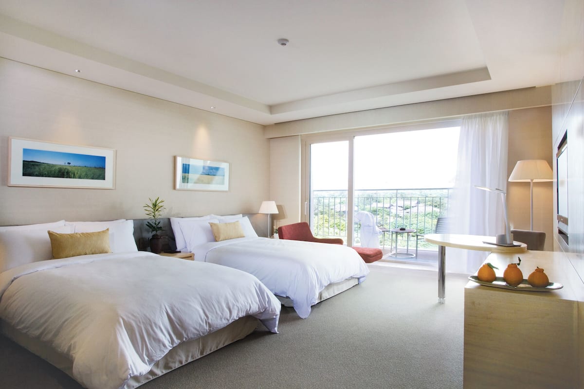 Une chambre d’hôtel avec deux lits, un balcon, un bureau avec une lampe et une vue sur l’extérieur.