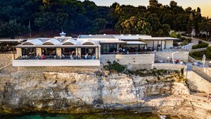 A coastal hotel with outdoor seating overlooking the sea, a stone facade, and a staircase leading to the upper levels. - Arena Verudela Beach Apartments (Pula)