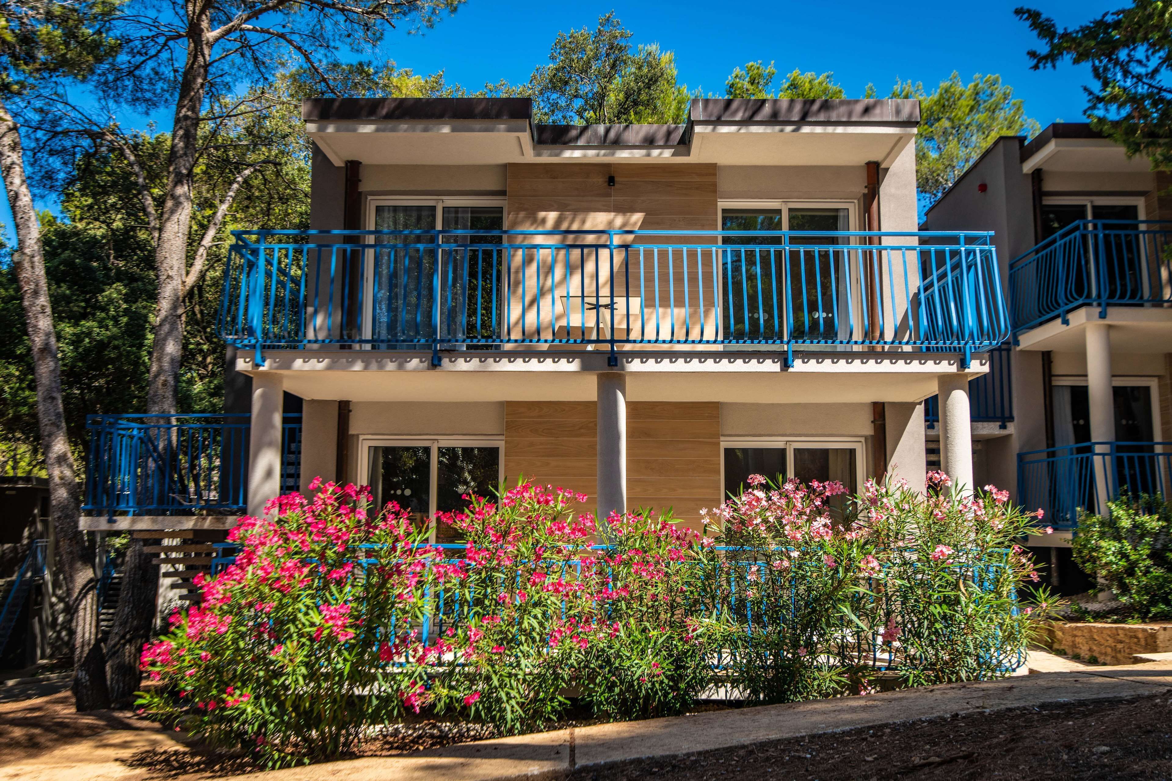 A two-story building with balconies and a garden with pink flowers.