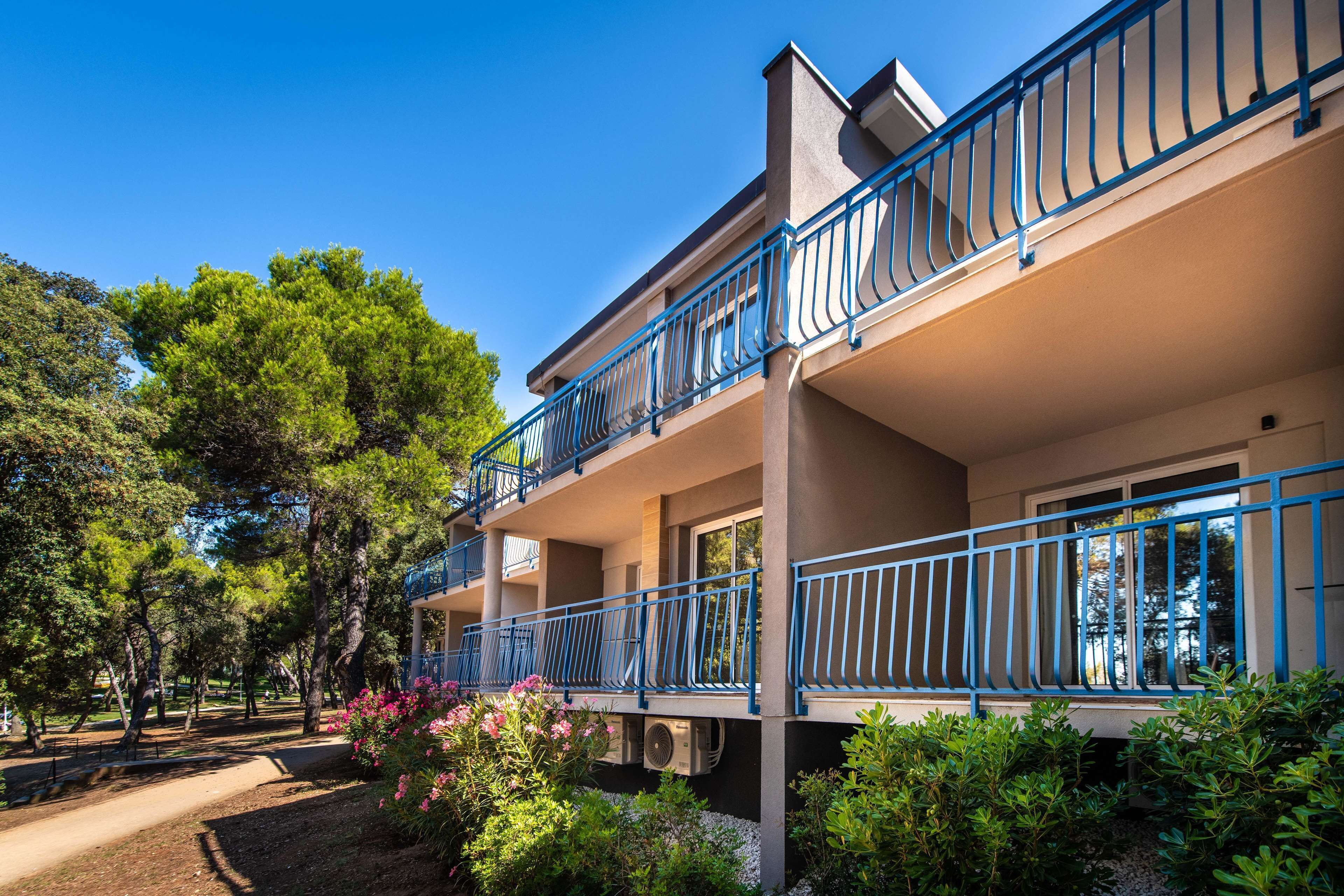 A two-story building with balconies, surrounded by trees and shrubs.