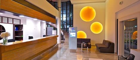 A modern hotel lobby with a reception desk, seating area, and a staircase leading to an upper level.