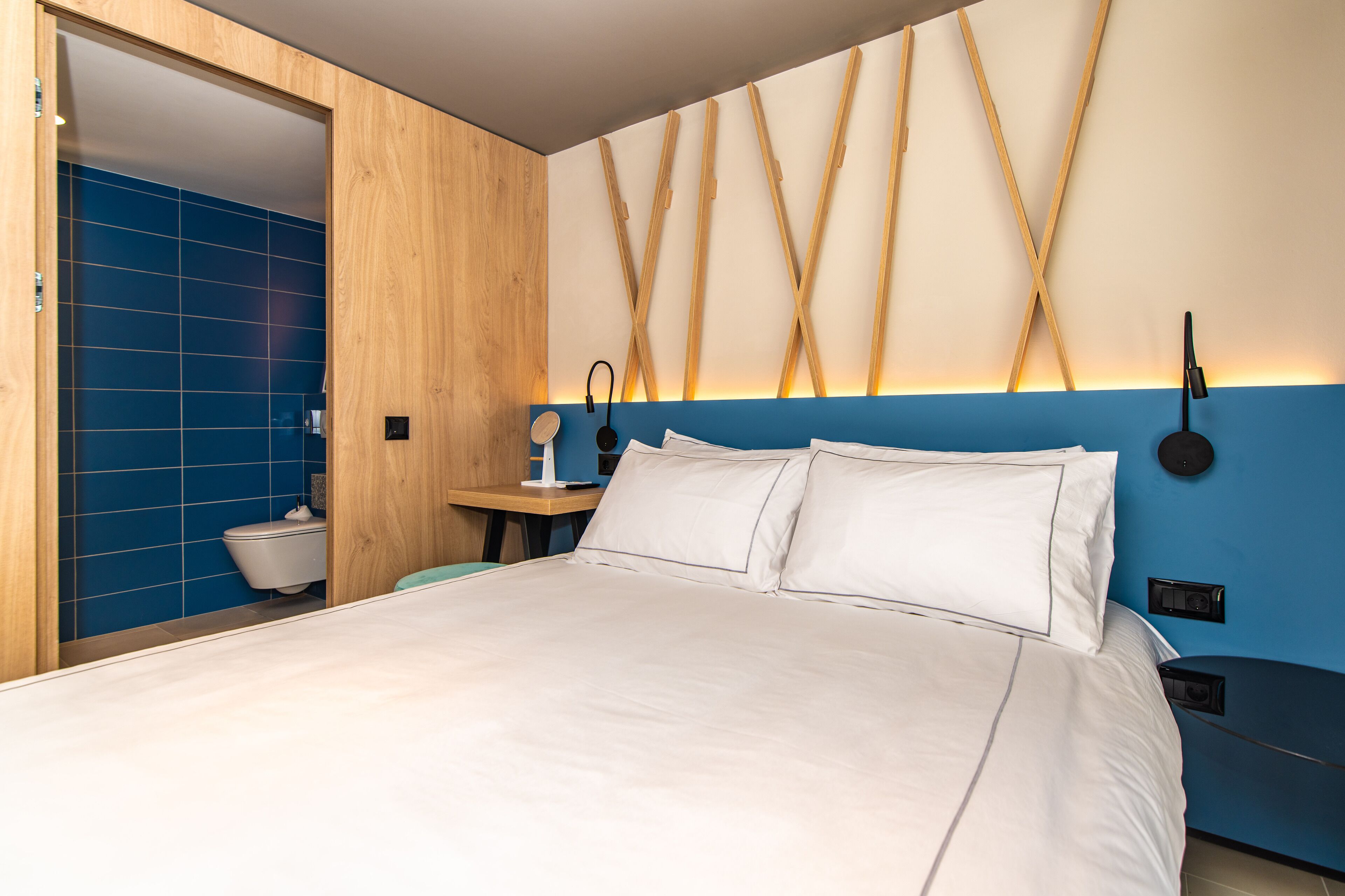 Een moderne hotelkamer met een groot bed, houten lambrisering, een blauwe accentmuur en een houten hoofdeinde met een bijzondere lamp.