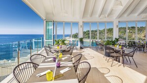 A modern outdoor dining area with tables and chairs, large windows overlooking the sea, and a view of the coastline. - Arena Verudela Beach Apartments (Pula)