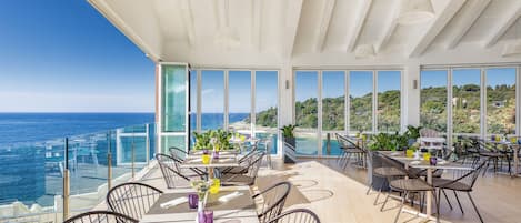 Un espacio moderno para comer al aire libre con mesas y sillas, amplias ventanas con vistas al mar y panorama de la costa.