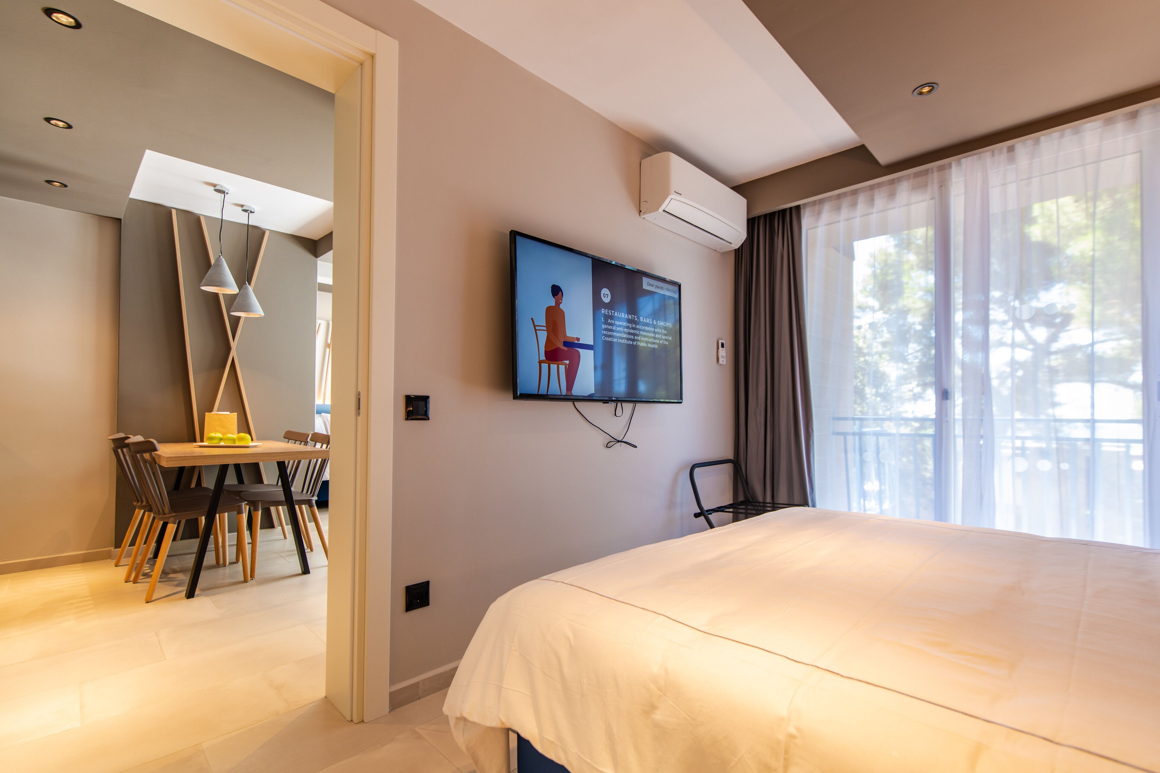 A hotel room with a bed, a desk with chairs, a television mounted on the wall, and a window with curtains.