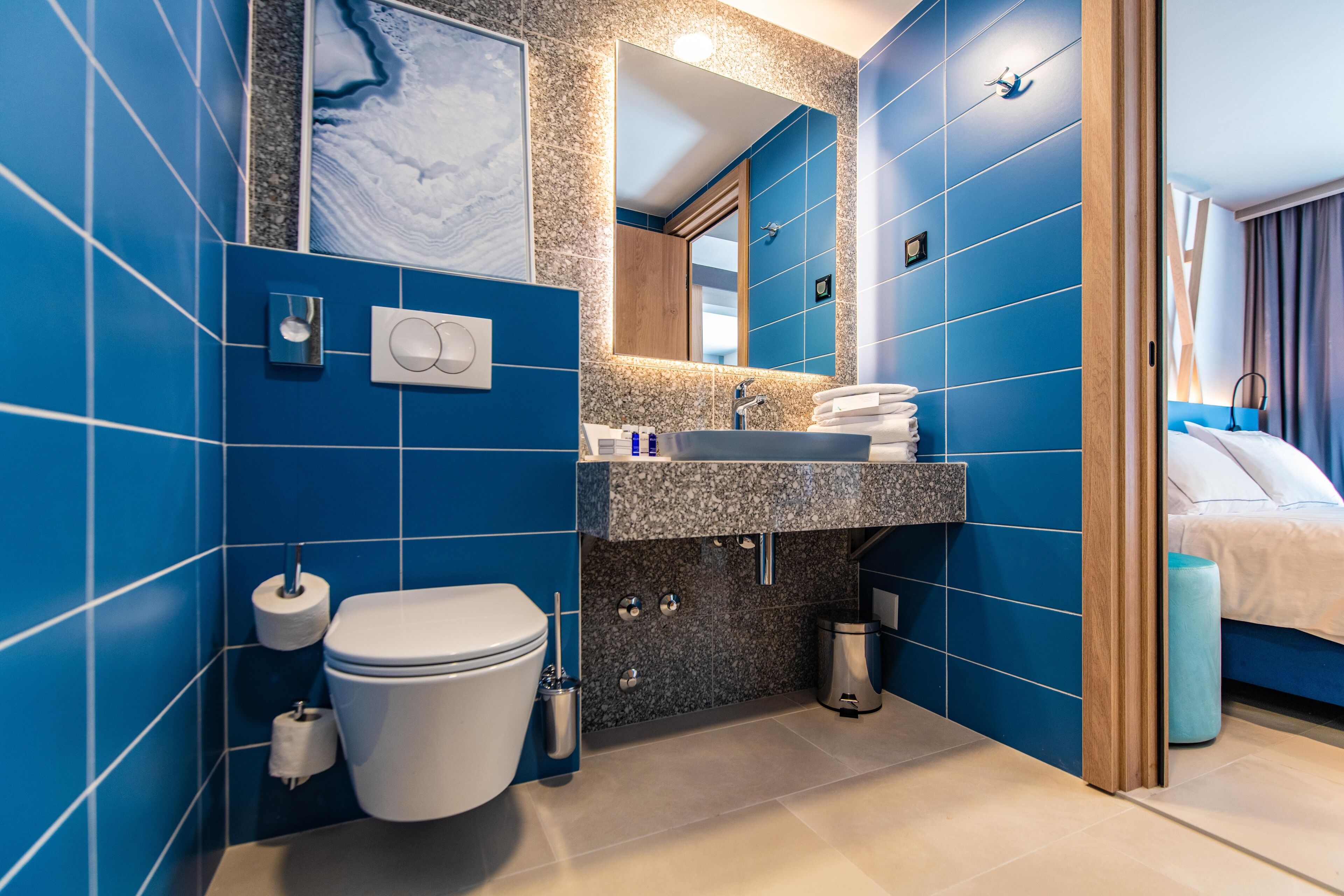 Ein modernes Badezimmer mit blauen Fliesen, einer weißen Toilette, einem Waschbecken mit Marmorplatte und einem Spiegel darüber.